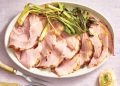 Sliced glazed ham served on a platter with roasted green onions and carving utensils nearby.