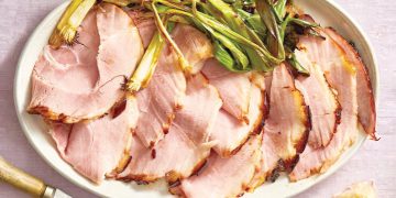 Sliced glazed ham served on a platter with roasted green onions and carving utensils nearby.