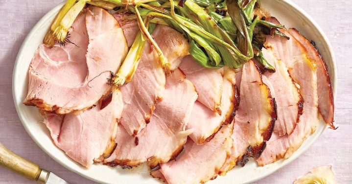 Sliced glazed ham served on a platter with roasted green onions and carving utensils nearby.