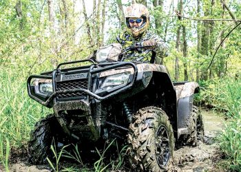 Rider in protective gear drives an ATV through a muddy trail in a wooded area.