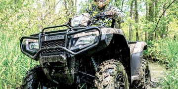 Rider in protective gear drives an ATV through a muddy trail in a wooded area.