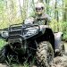 Rider in protective gear drives an ATV through a muddy trail in a wooded area.