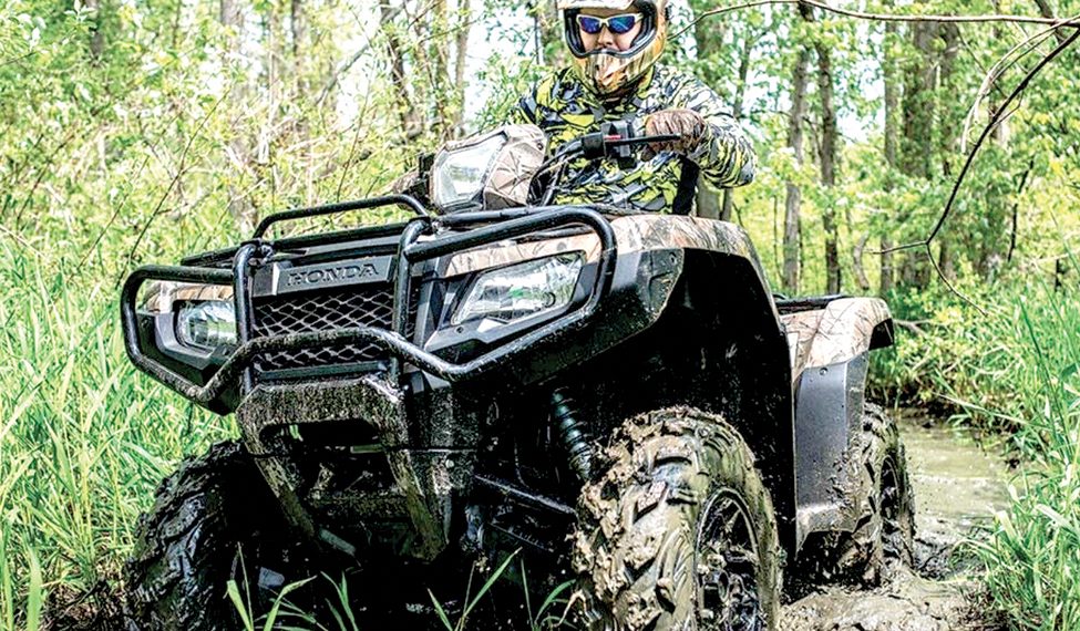Rider in protective gear drives an ATV through a muddy trail in a wooded area.