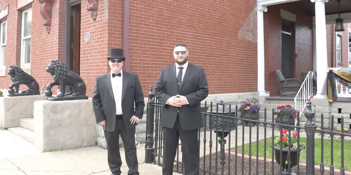 Two men in formal suits stand outside a historic brick building with decorative details and lion statues.