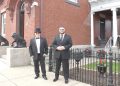 Two men in formal suits stand outside a historic brick building with decorative details and lion statues.