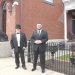 Two men in formal suits stand outside a historic brick building with decorative details and lion statues.