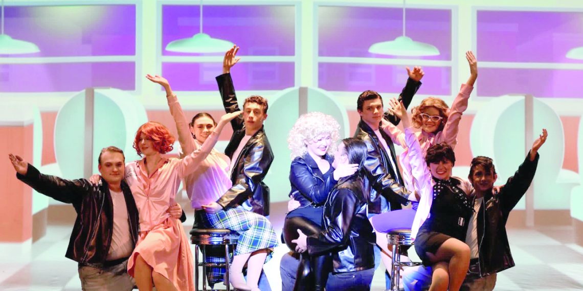 Cast of a Grease-themed musical in 1950s-style costumes posing on a diner-themed stage with bright lights and raised arms.