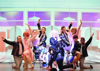 Cast of a Grease-themed musical in 1950s-style costumes posing on a diner-themed stage with bright lights and raised arms.