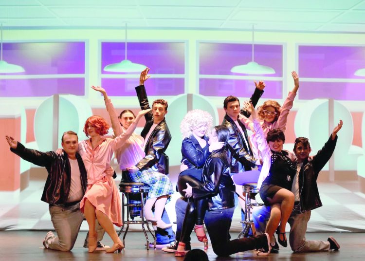 Cast of a Grease-themed musical in 1950s-style costumes posing on a diner-themed stage with bright lights and raised arms.