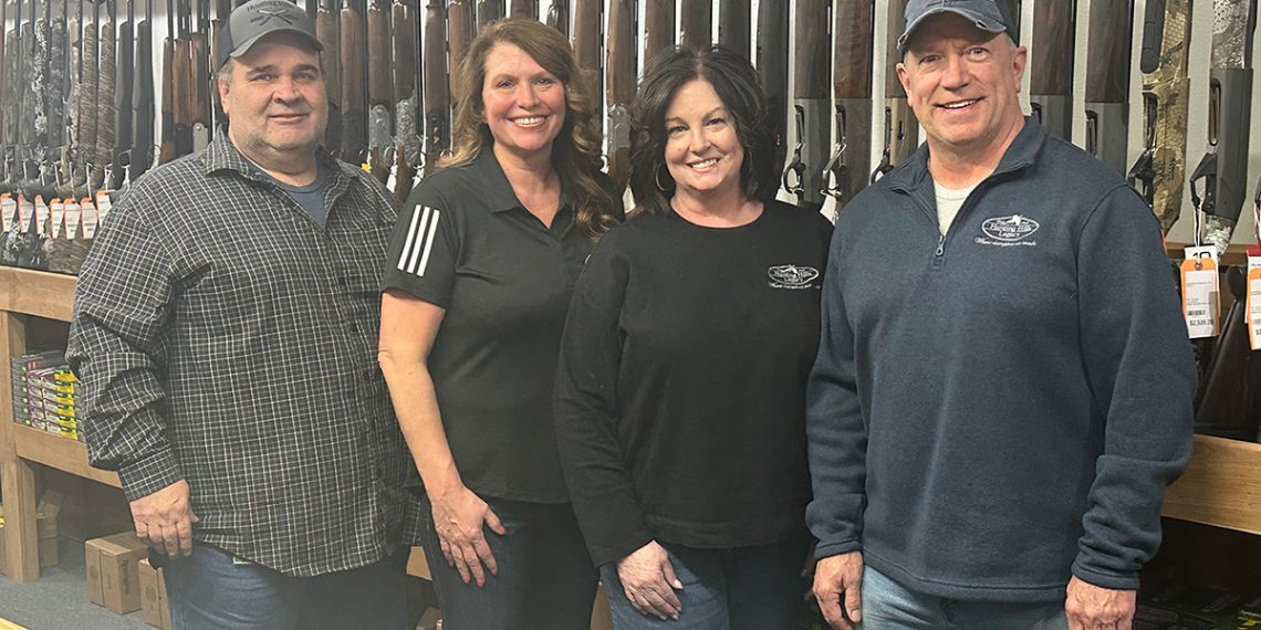 Four people pose inside a gun shop with rifles and shooting supplies displayed behind them.