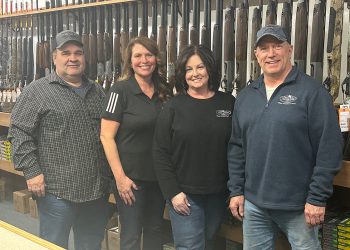Four people pose inside a gun shop with rifles and shooting supplies displayed behind them.
