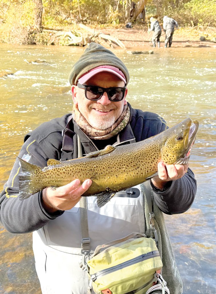 Man holding a large trout while standing in a stream with other people fishing in the background.