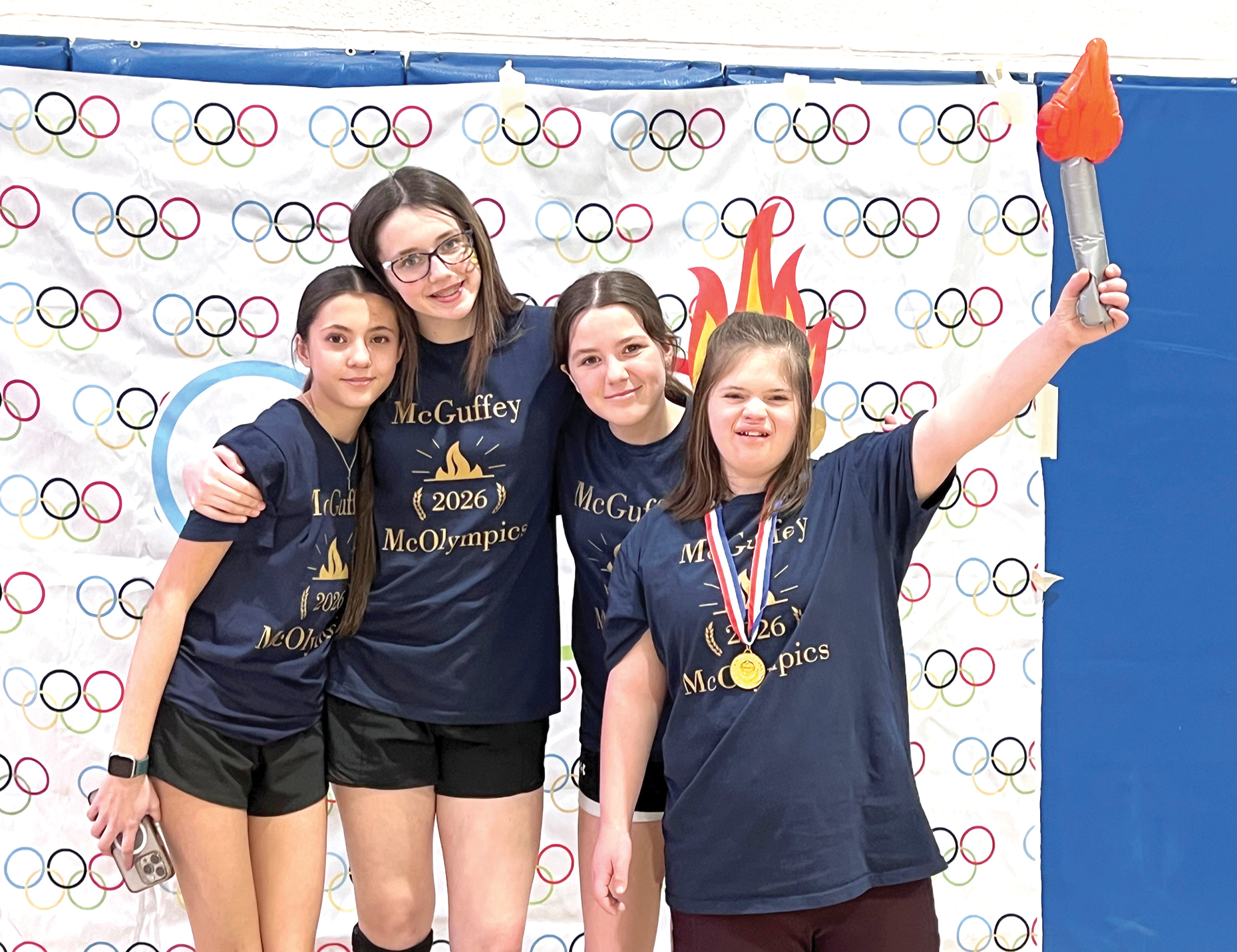 Four students pose with a medal and torch prop in front of an Olympic-themed backdrop at McOlympics.