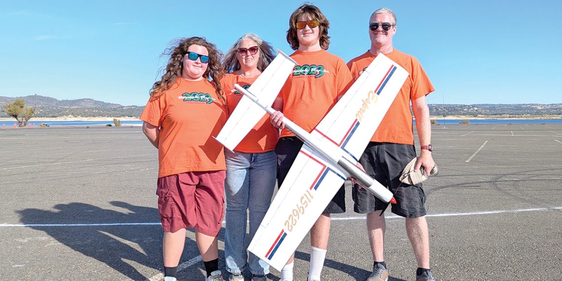 Four people in matching shirts pose with a large model airplane outdoors on a sunny day.