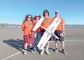Four people in matching shirts pose with a large model airplane outdoors on a sunny day.