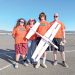 Four people in matching shirts pose with a large model airplane outdoors on a sunny day.
