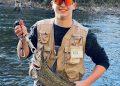 Young angler holds a trout while standing in a stream with another fisherman in the background.