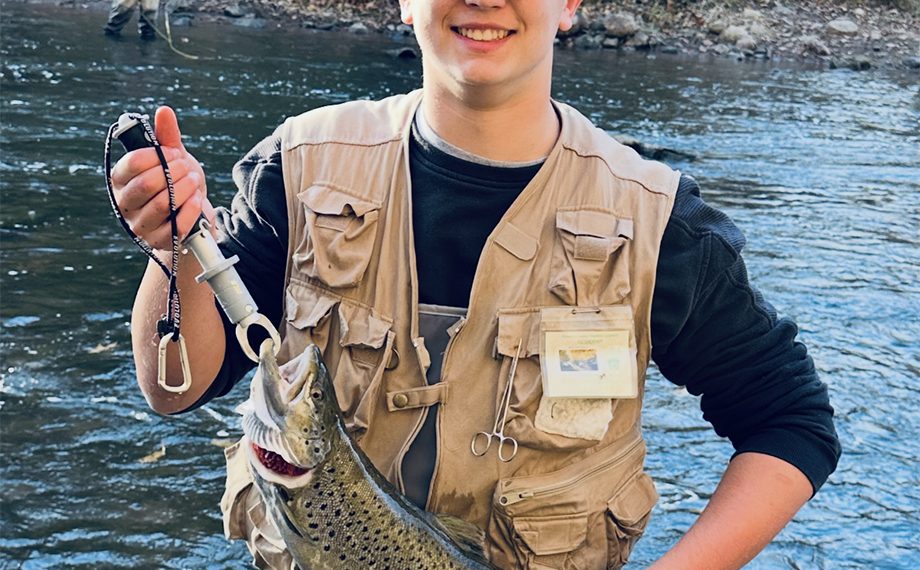 Young angler holds a trout while standing in a stream with another fisherman in the background.