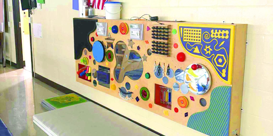 Colorful sensory activity board with interactive elements mounted on a school hallway wall.