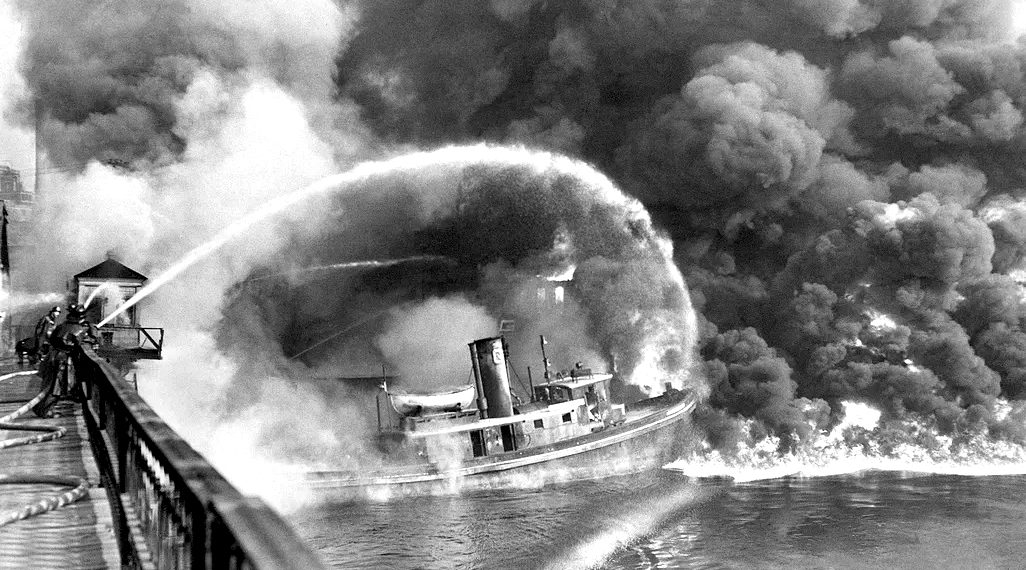 Firefighters spraying water onto a burning boat with heavy smoke rising over the water.