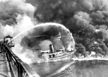 Firefighters spraying water onto a burning boat with heavy smoke rising over the water.