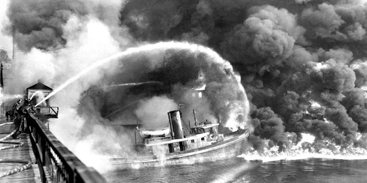 Firefighters spraying water onto a burning boat with heavy smoke rising over the water.
