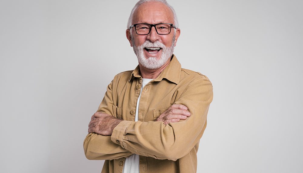 Smiling older man with glasses and gray beard standing with arms crossed against a plain background.