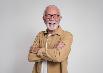 Smiling older man with glasses and gray beard standing with arms crossed against a plain background.