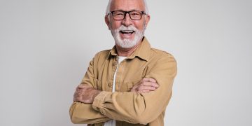 Smiling older man with glasses and gray beard standing with arms crossed against a plain background.