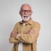 Smiling older man with glasses and gray beard standing with arms crossed against a plain background.