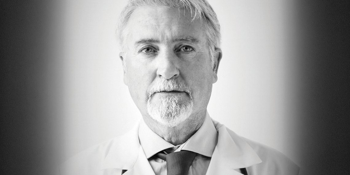 Black and white portrait of an older doctor wearing a white coat and looking at the camera.
