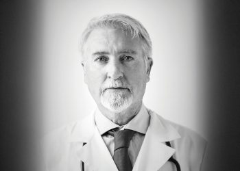 Black and white portrait of an older doctor wearing a white coat and looking at the camera.