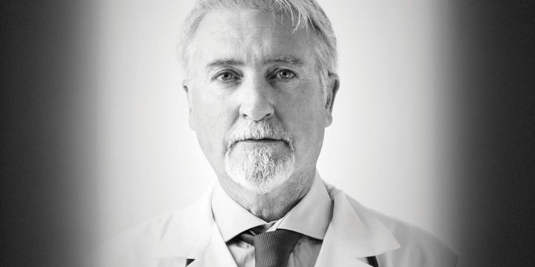 Black and white portrait of an older doctor wearing a white coat and looking at the camera.