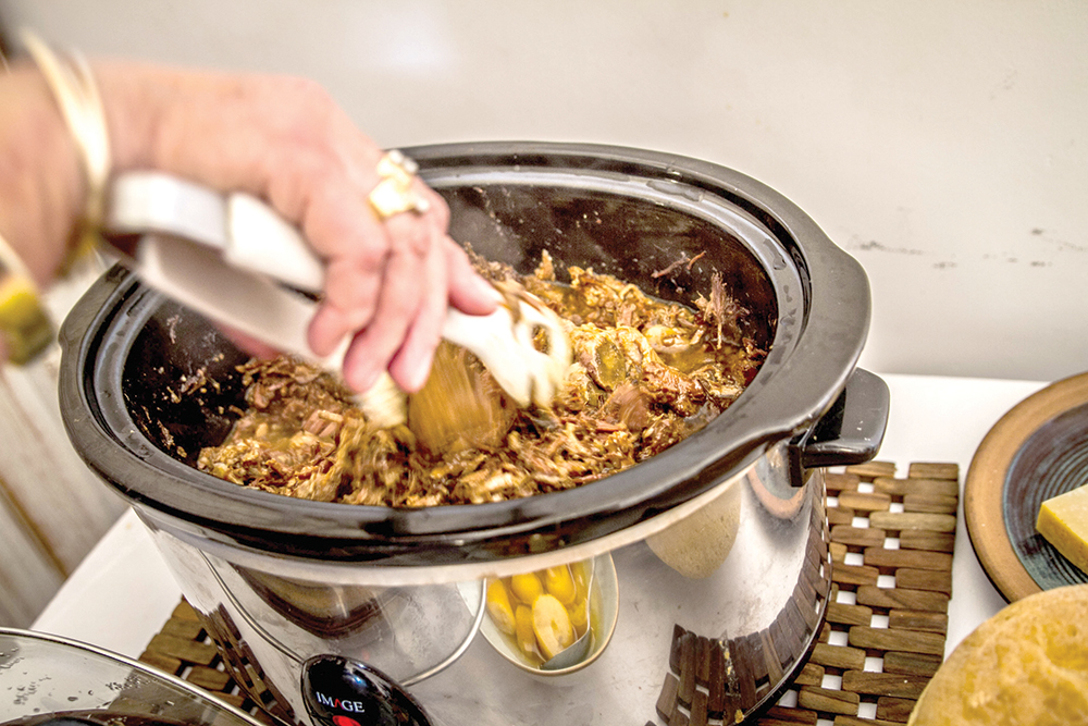 Person using tongs to stir shredded meat cooking in a slow cooker on a kitchen counter.