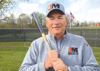 A baseball coach in a Jefferson-Morgan Rockets jacket holds a bat while standing on a field.