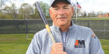 A baseball coach in a Jefferson-Morgan Rockets jacket holds a bat while standing on a field.