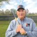 A baseball coach in a Jefferson-Morgan Rockets jacket holds a bat while standing on a field.