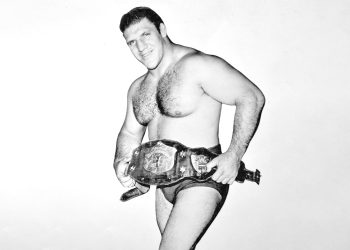A black and white image of a professional wrestler holding a championship belt and posing confidently.