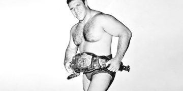 A black and white image of a professional wrestler holding a championship belt and posing confidently.