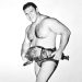 A black and white image of a professional wrestler holding a championship belt and posing confidently.