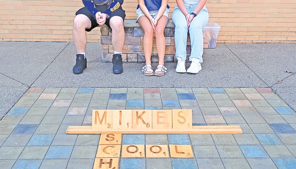 Three students sit on a bench behind a large Scrabble-style display spelling “Mikes School” on the ground.