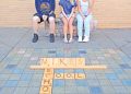 Three students sit on a bench behind a large Scrabble-style display spelling “Mikes School” on the ground.
