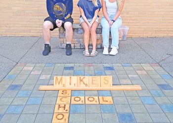 Three students sit on a bench behind a large Scrabble-style display spelling “Mikes School” on the ground.