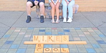 Three students sit on a bench behind a large Scrabble-style display spelling “Mikes School” on the ground.