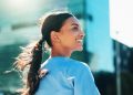Smiling woman with a ponytail looking back outdoors with city buildings in the background.