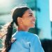 Smiling woman with a ponytail looking back outdoors with city buildings in the background.