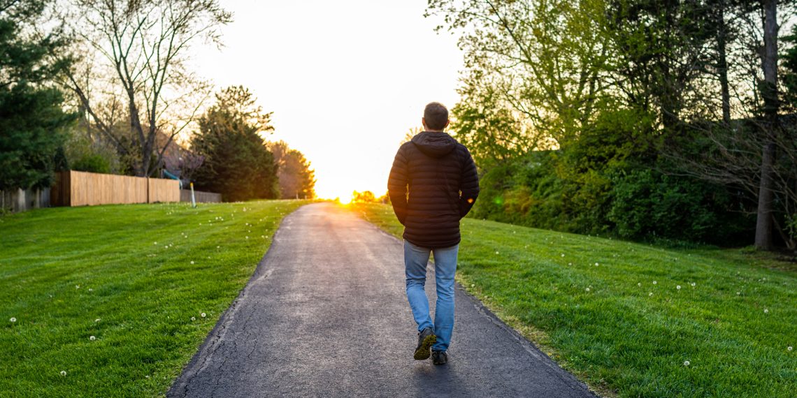 Person walking down a path toward a sunset with trees and grass on both sides.