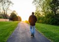 Person walking down a path toward a sunset with trees and grass on both sides.