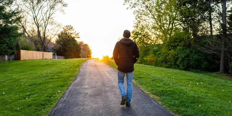 Person walking down a path toward a sunset with trees and grass on both sides.