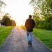 Person walking down a path toward a sunset with trees and grass on both sides.